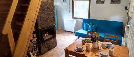 Une pièce chaleureuse avec une cheminée en pierre, une table à manger en bois mise avec des assiettes et des tasses, un canapé bleu agrémenté de coussins et une fenêtre offrant une vue agréable.