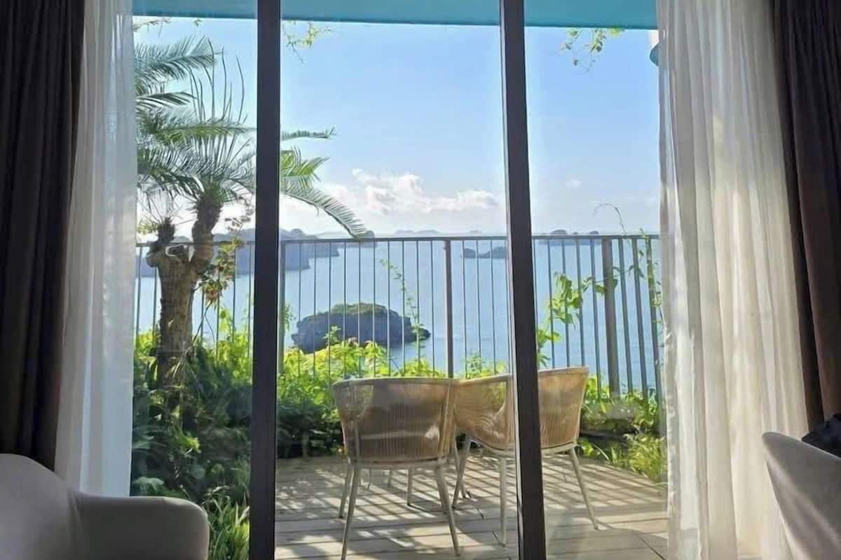 A room with a large sliding glass door leading to an outdoor balcony with a view of the sea and a palm tree. There are two chairs and a small round table on the balcony.