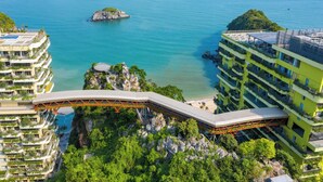 A modern hotel with a unique curved bridge connecting two buildings, overlooking a coastal area with clear blue waters and rocky islands.