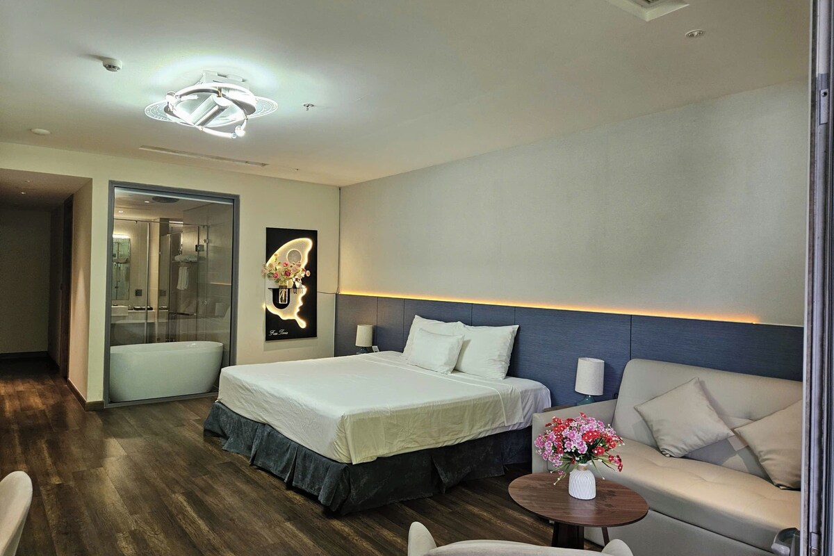 A modern hotel room with a large bed, a sofa, a small table with flowers, and a unique wall art piece.