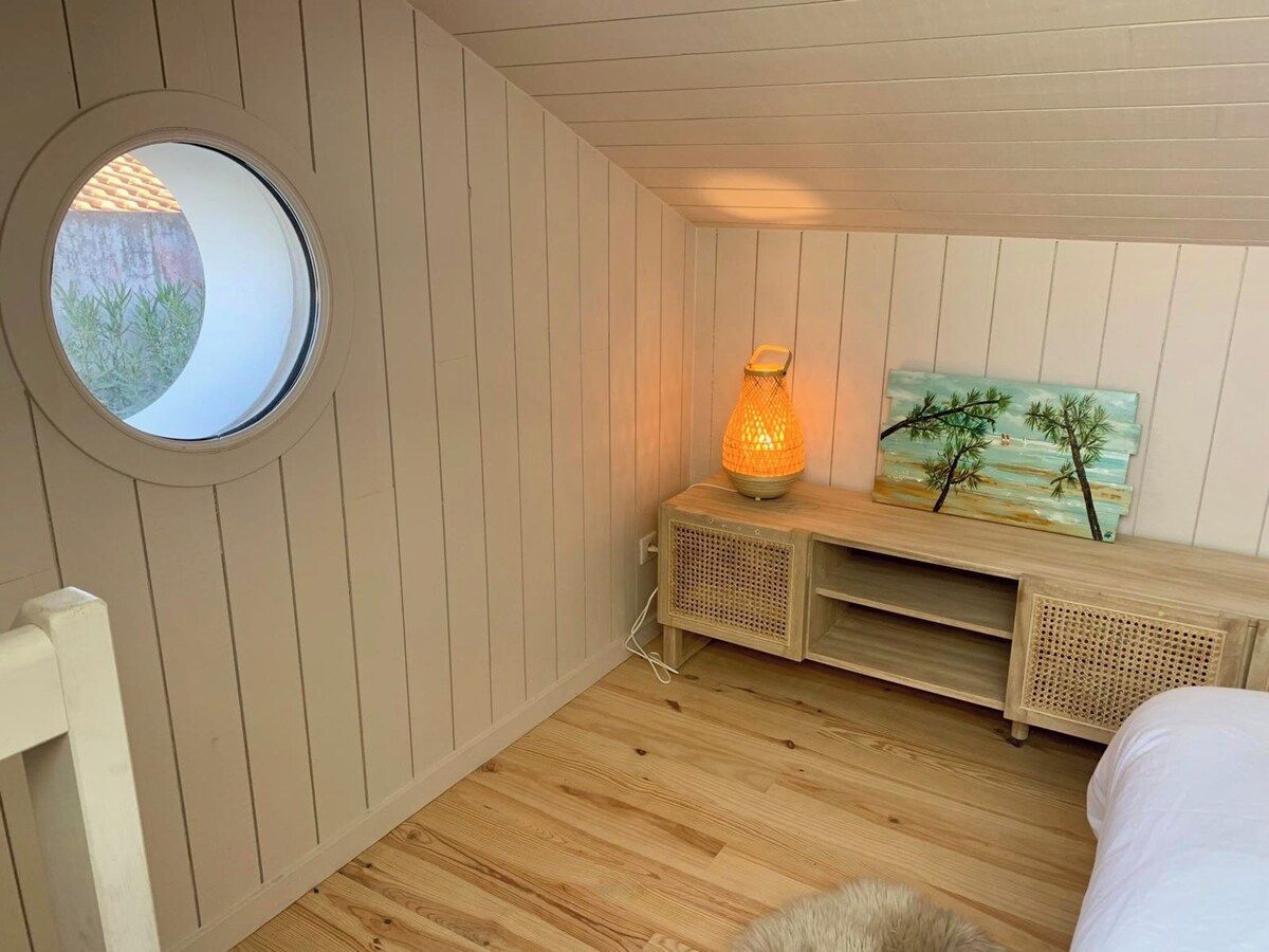 Une pièce avec une fenêtre ronde, un lit, une table de chevet avec une lampe et une étagère fixée au mur, sur laquelle est posée une peinture.