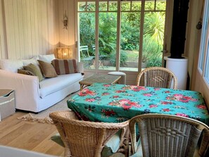 Une pièce chaleureuse avec un canapé blanc, une table recouverte d’une nappe à motifs floraux, des chaises en osier et une vue sur la verdure à l’extérieur.