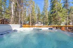 A hot tub surrounded by tall trees, with a wooden deck and a house in the background. - Donner Lake Outpost: Hot Tub, Game Room, Walk to Beach (Truckee)