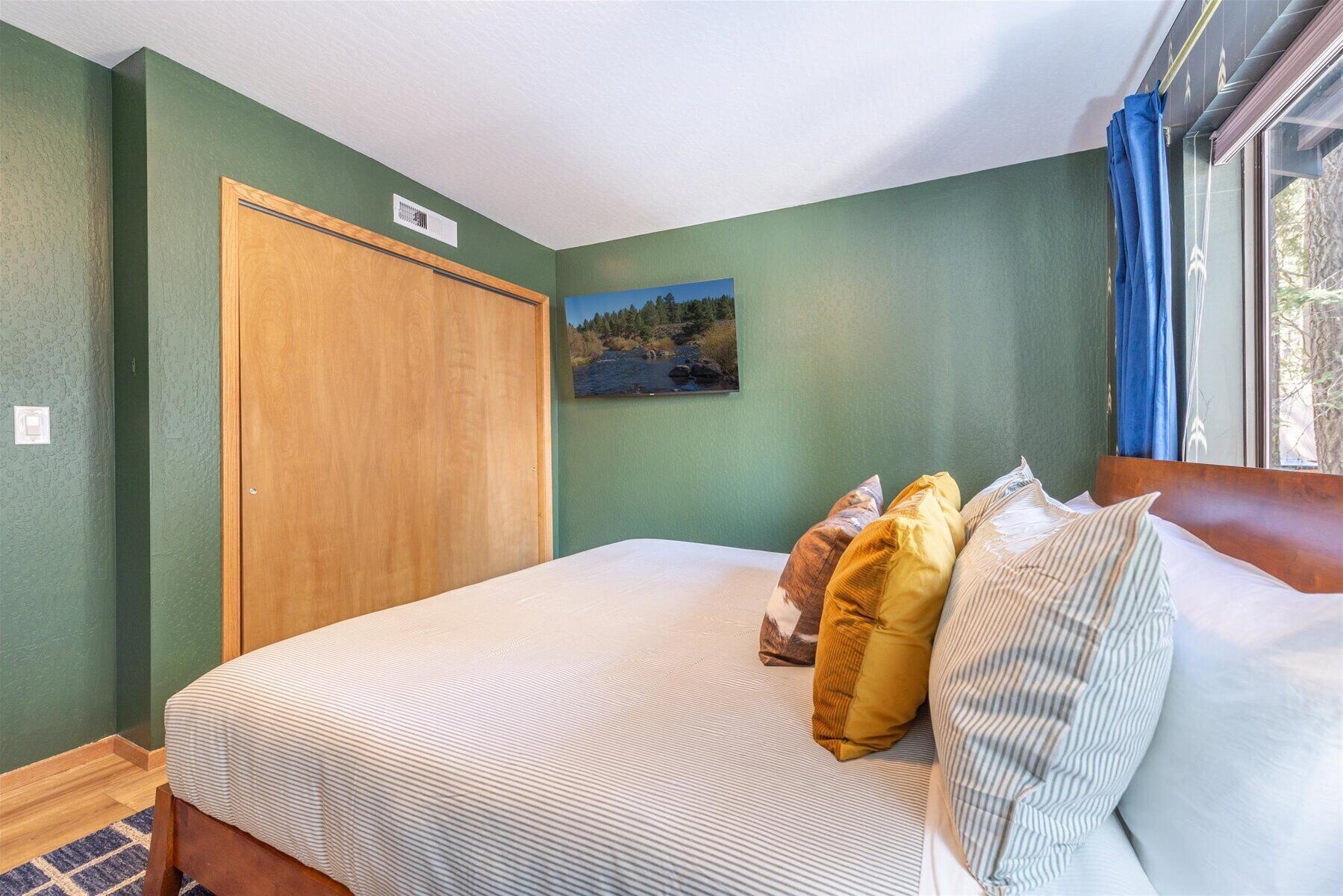 Une chambre à coucher avec un lit, des oreillers, un tableau, une porte en bois et une fenêtre avec des rideaux.