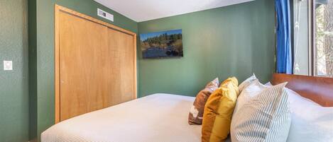 Une chambre à coucher avec un lit, des oreillers, un tableau, une porte en bois et une fenêtre avec des rideaux.