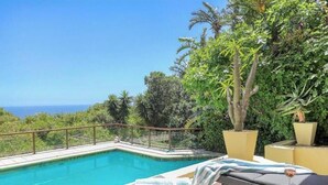 Ein Pool mit freiem Blick auf den Ozean, umgeben von üppiger Vegetation und einem Glasgeländer.