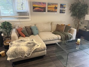 A living room with a sofa, a glass coffee table, a potted plant, and wall art. - Peaceful Cottage for 2 – Fire Pit, BBQ & Nature! (Norland)
