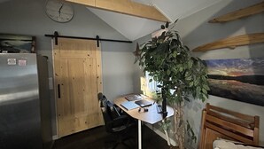 Una habitació amb una porta de fusta, un escriptori amb una cadira, una planta, un rellotge a la paret i un quadre.
