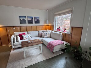 Ett vardagsrum med en vit soffa, ett träbord, en mönstrad pläd och ett fönster med dekorativa föremål.