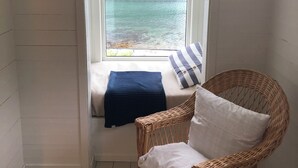 A room with a window seat overlooking a body of water, a wicker chair, and a white wall.