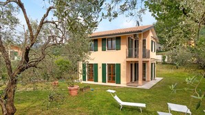 Uma casa de dois andares com uma varanda, um gramado com espreguiçadeiras e um vaso de planta.