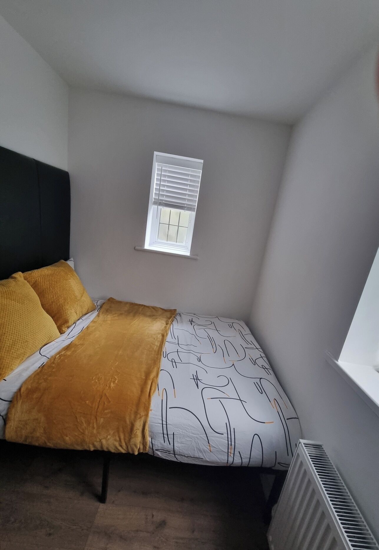 A small, single-bed room with a window, a radiator, and a headboard.
