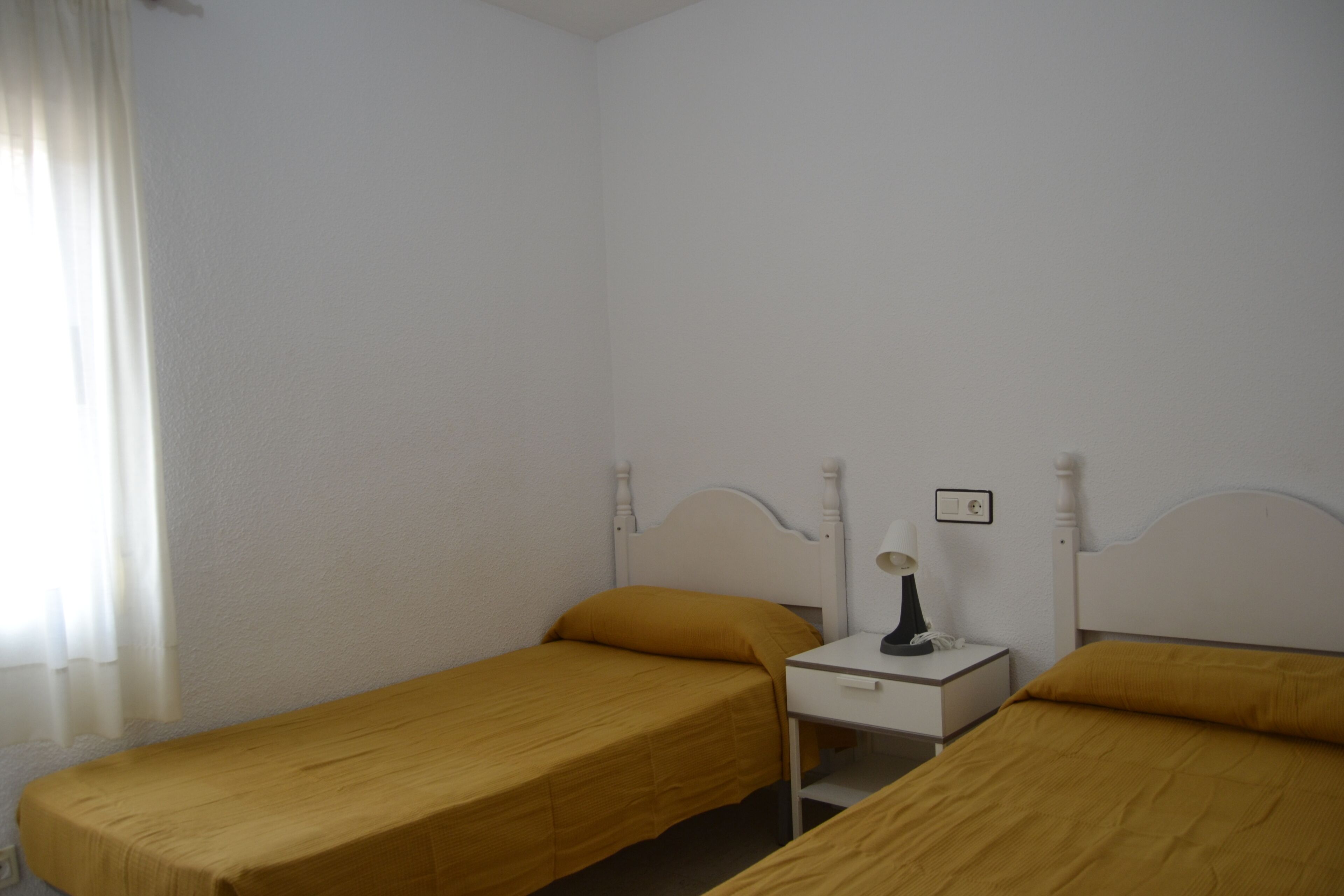 Una habitación pequeña individual con dos camas, una mesita de noche y una ventana con cortinas.