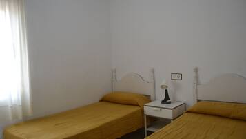 Una habitación pequeña individual con dos camas, una mesita de noche y una ventana con cortinas.