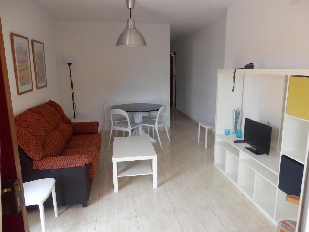 Una sala de estar con un sofá, una mesa de comedor con sillas, un televisor sobre un mueble bajo y un estante blanco.