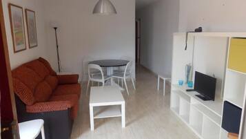 Una sala de estar con un sofá, una mesa de comedor con sillas, un televisor sobre un mueble bajo y un estante blanco.