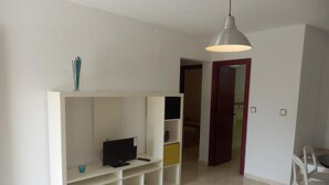 Une pièce avec un téléviseur posé sur un meuble blanc, une table à manger avec des chaises, et une porte rouge à l’arrière-plan.
