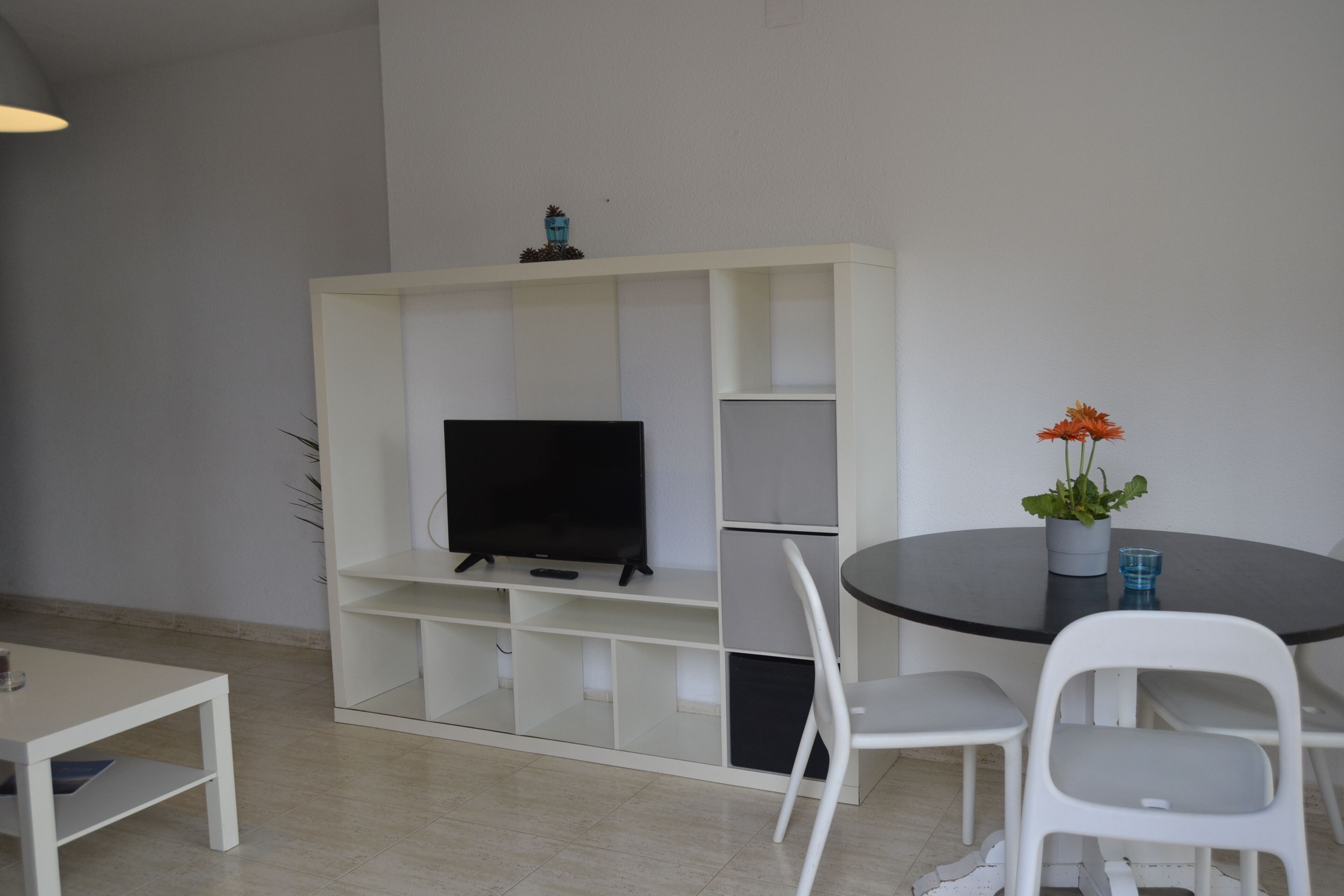 Una habitación con un mueble blanco para televisor, una mesa redonda con sillas y una planta en maceta.