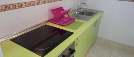 A kitchen with a green countertop, a black induction cooktop, a pink dish rack, a stainless steel sink, and a microwave.