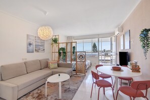 Ein modernes Wohnzimmer mit einer Couch, einem runden Tisch mit Stühlen, einem kleinen weißen Tisch und einem großen Fenster.