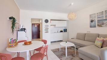 A modern living room with a sofa, a dining table with chairs, a kitchen area, and a hallway.