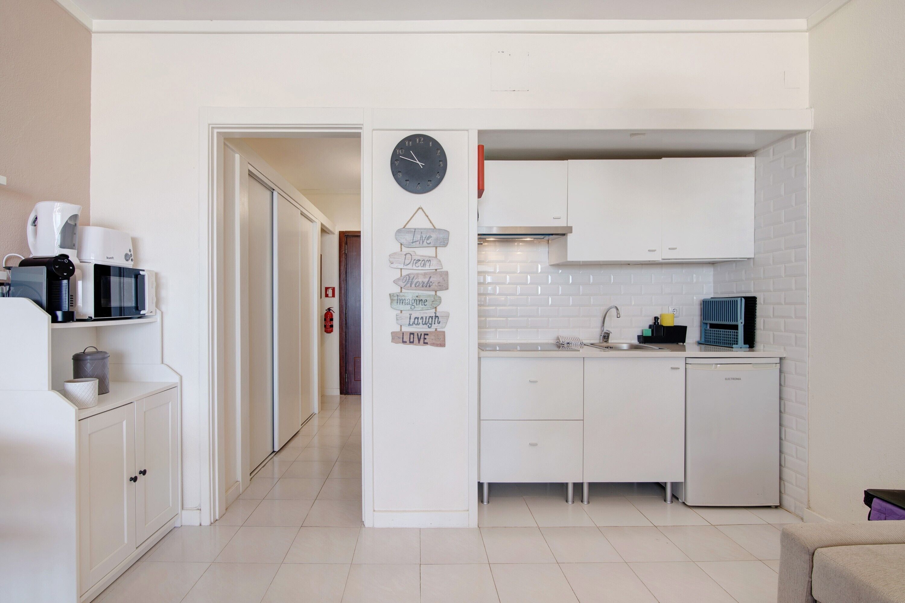 Une cuisine de taille réduite avec des meubles blancs, un four à micro-ondes et une horloge au mur.