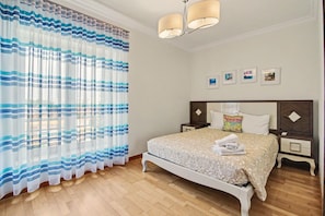 A bedroom with a bed, nightstands, a chandelier, and a window with curtains. - Holiday Home 'Brisa Do Mar Moura Beach' with Shared Pool, Shared Garden and Wi-Fi (Quarteira)