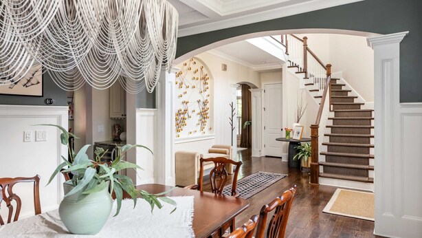 Un comedor con mesa de madera, sillas, un florero con planta, una araña y una escalera que lleva a otra habitación.