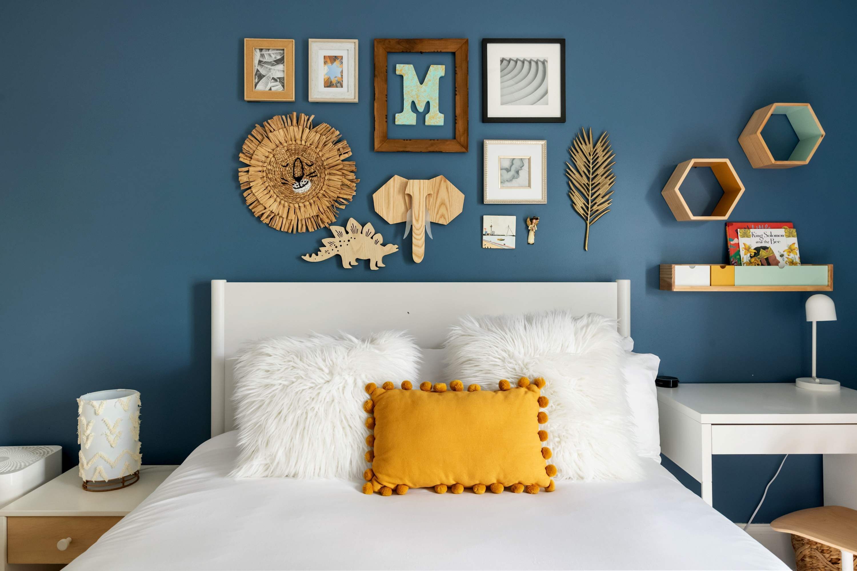 A bedroom with a bed, white fluffy pillows, a mustard-colored pillow, a nightstand with a lamp, and wall decorations including framed pictures, a wooden 'M', and a sunburst design.