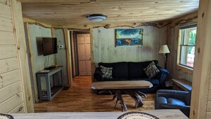 A wooden cabin interior with a wooden table, a sofa, a TV, and a painting on the wall. - RiverFront Cabin in New River Gorge National Park & Preserve (Prince)