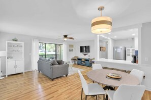 Ein modernes Wohnzimmer mit grauem Sofa, einem Esstisch mit Stühlen, einem Deckenventilator und einer Küchenzeile mit Kühlschrank und Fernseher.