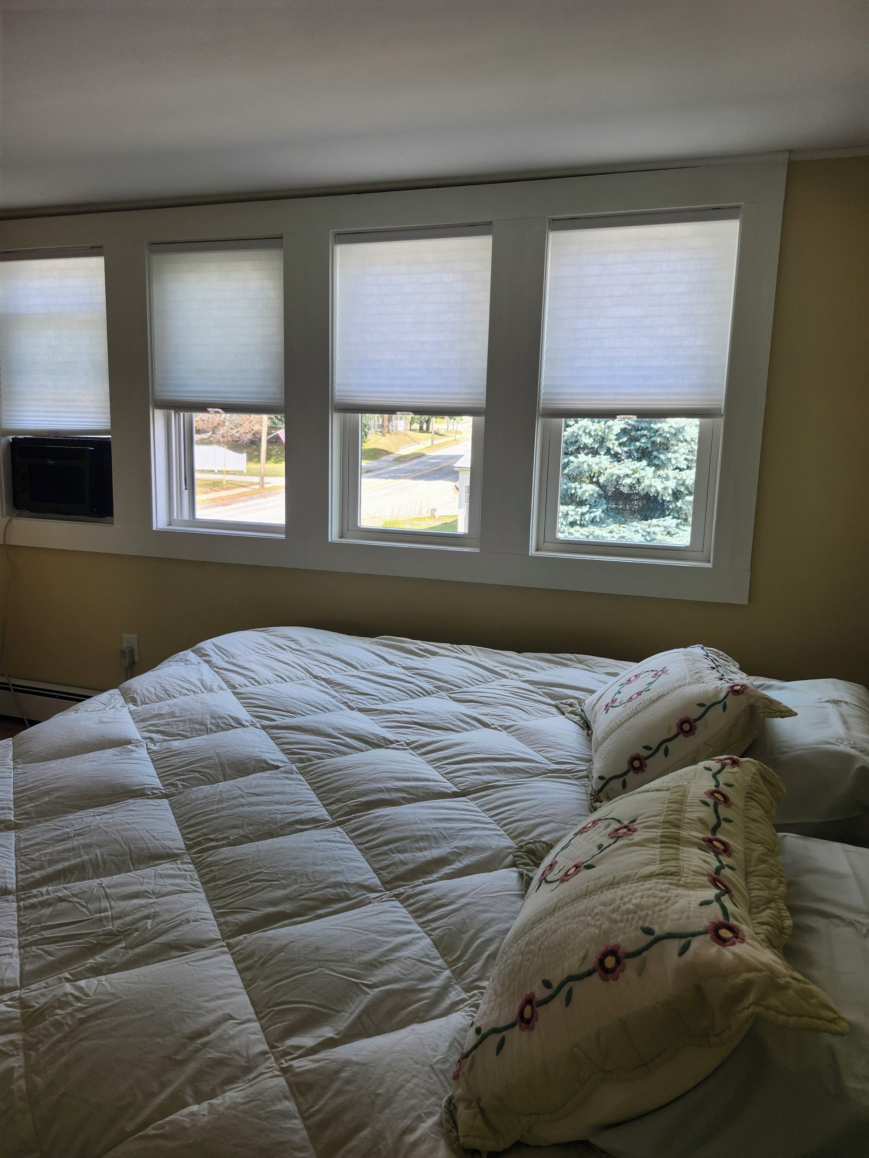 Un dormitorio con una cama grande, tres ventanas con persianas y vistas a la calle y árboles.