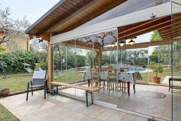 A patio with a table and chairs, a bench, and a view of a swimming pool and garden.