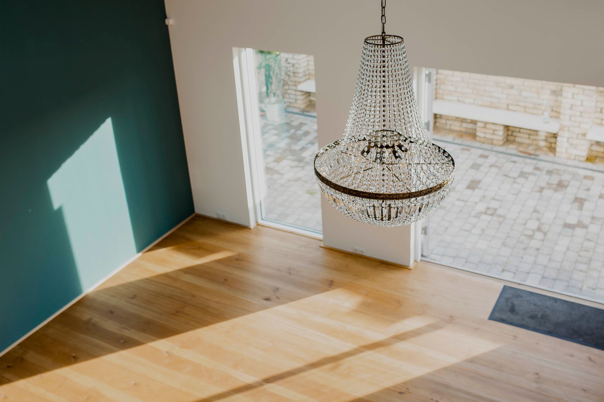A chandelier in a room with wooden flooring and a view of a brick wall through a window.