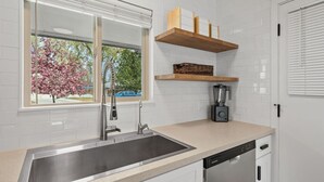 A modern kitchen with a stainless steel sink, a built-in dishwasher, a window with a view of trees, and a wooden shelf above the counter.