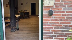 Een houten interieur met een bakstenen muur, een bordje met de tekst 'Klosterhof 16' en een deur die leidt naar een kamer met een tafel en stoelen.