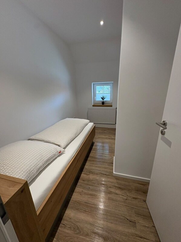 Ein schmaler Raum mit einem Einzelbett, einem Heizkörper, einem Fenster mit Jalousien und einer Tür.