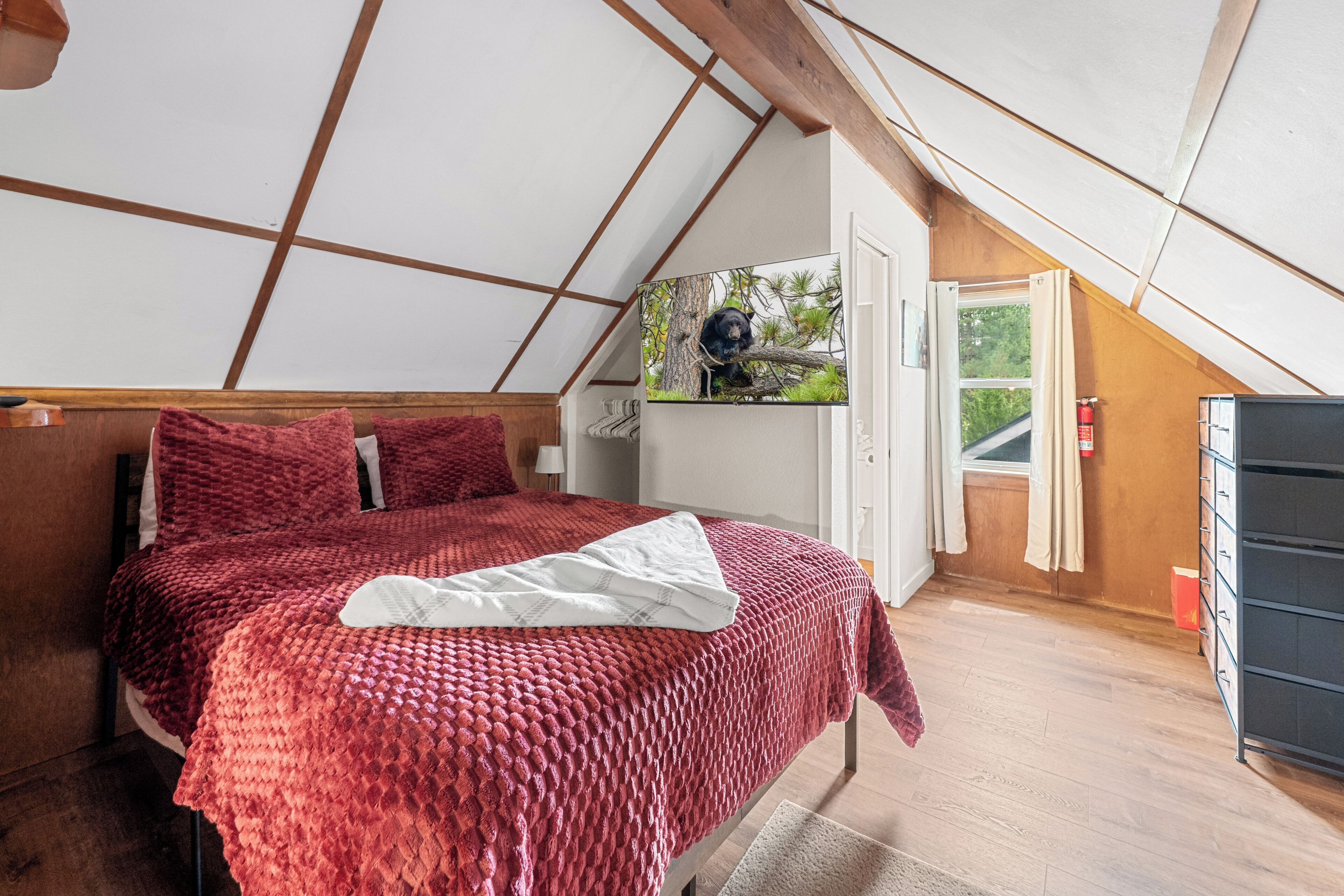 A bedroom with a sloped ceiling, a bed with red bedding, a window with a view of greenery, and a fire extinguisher near the window.