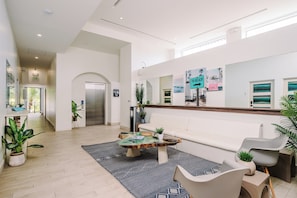 A modern hotel lobby with a reception desk, a seating area with a sofa and chairs, a coffee table, and a large mirror reflecting artwork and a beach scene. - Iguana Surf Boutique Hotel Tamarindo (Tamarindo)