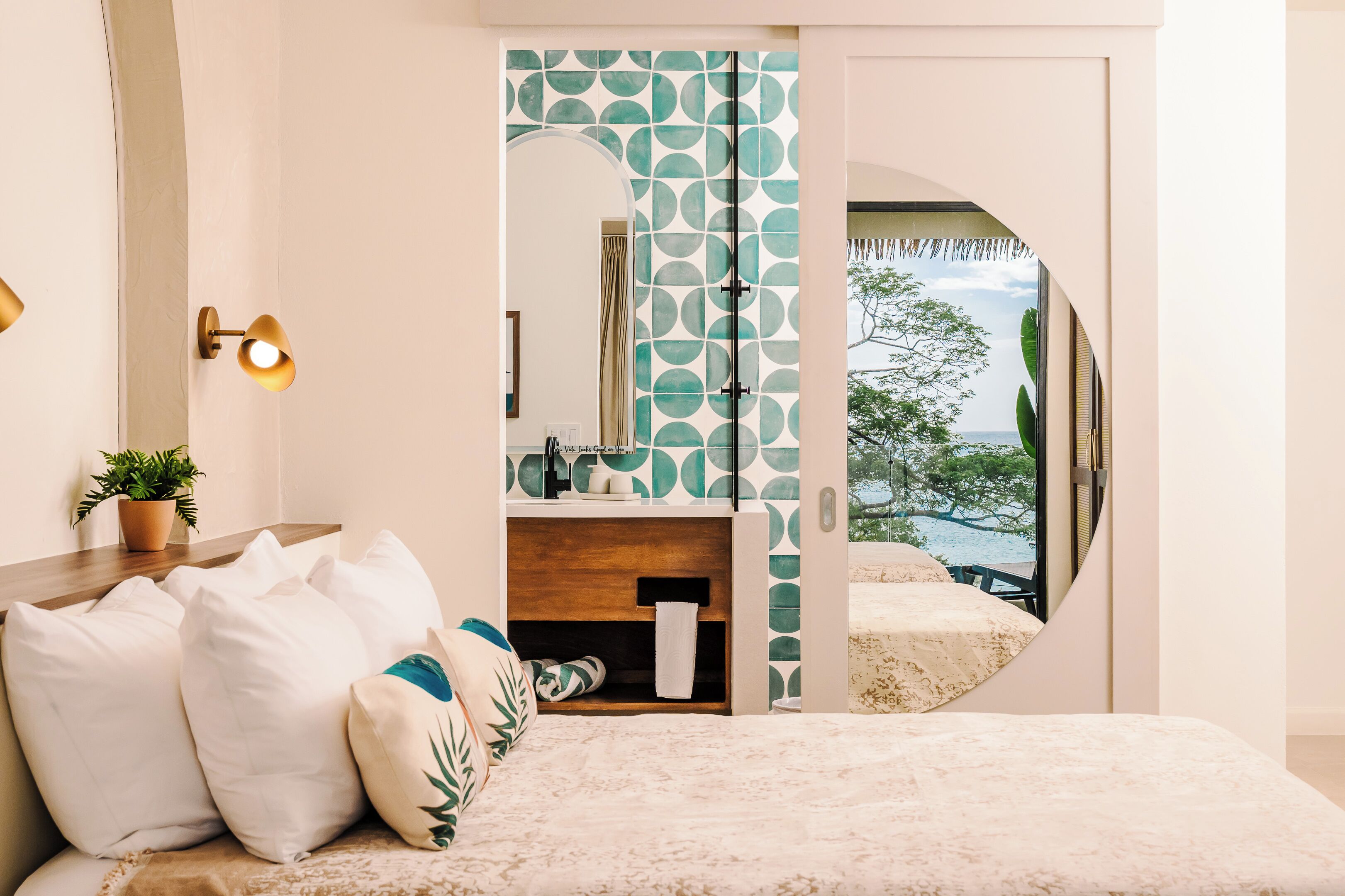 Une chambre à coucher avec un lit, des oreillers, une table de chevet, une plante en pot, un miroir et une vue sur l’extérieur.