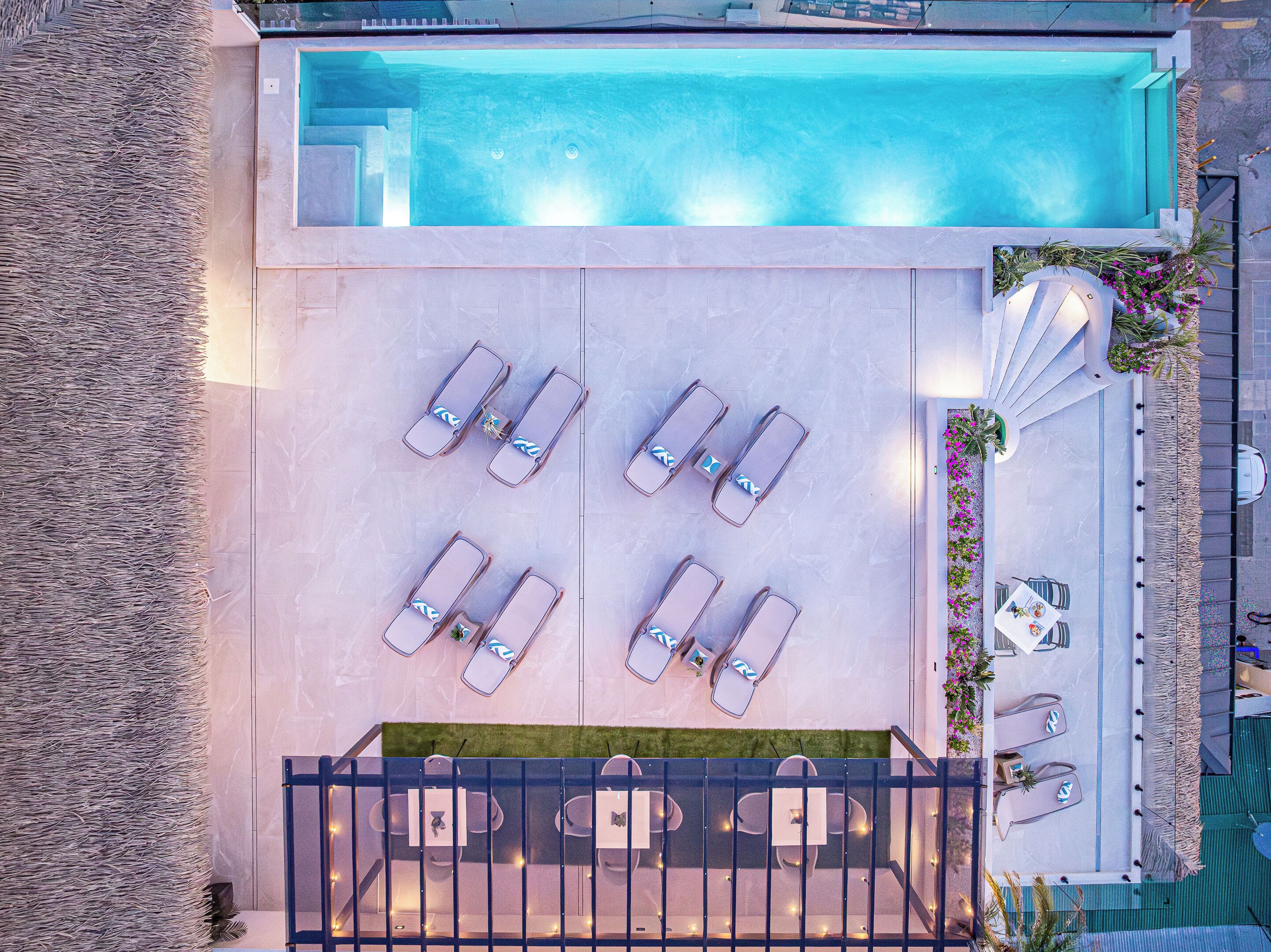 An aerial view of a pool area with lounge chairs arranged in two rows, a dining table with chairs, and a small garden area with flowers and plants.