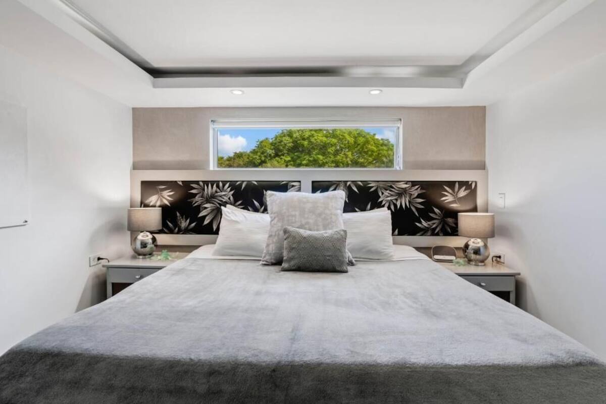 A modern bedroom with a large bed, two bedside tables, a window with a view of greenery, and a ceiling light.