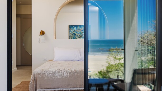 A bedroom with a bed, a painting, and a view of the beach through a glass door. - Iguana Surf Boutique Hotel Tamarindo (Tamarindo)