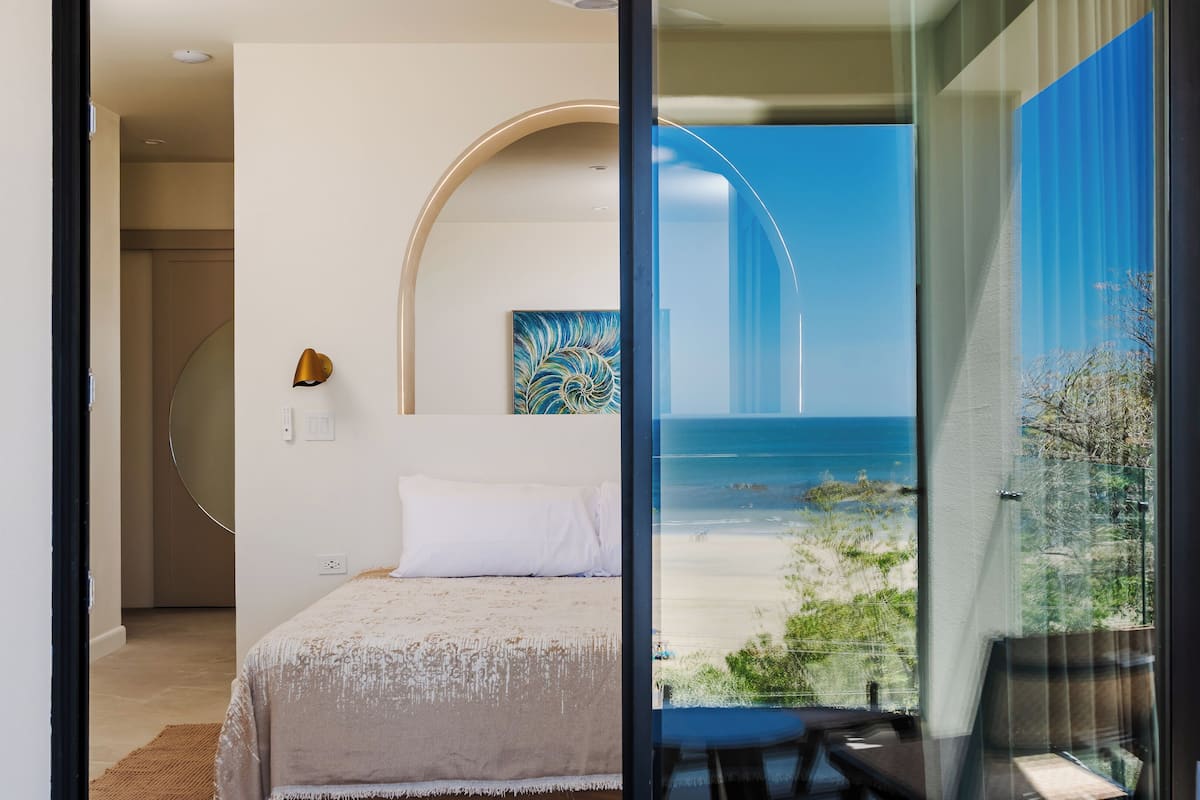 A bedroom with a bed, a painting, and a view of the beach through a glass door.