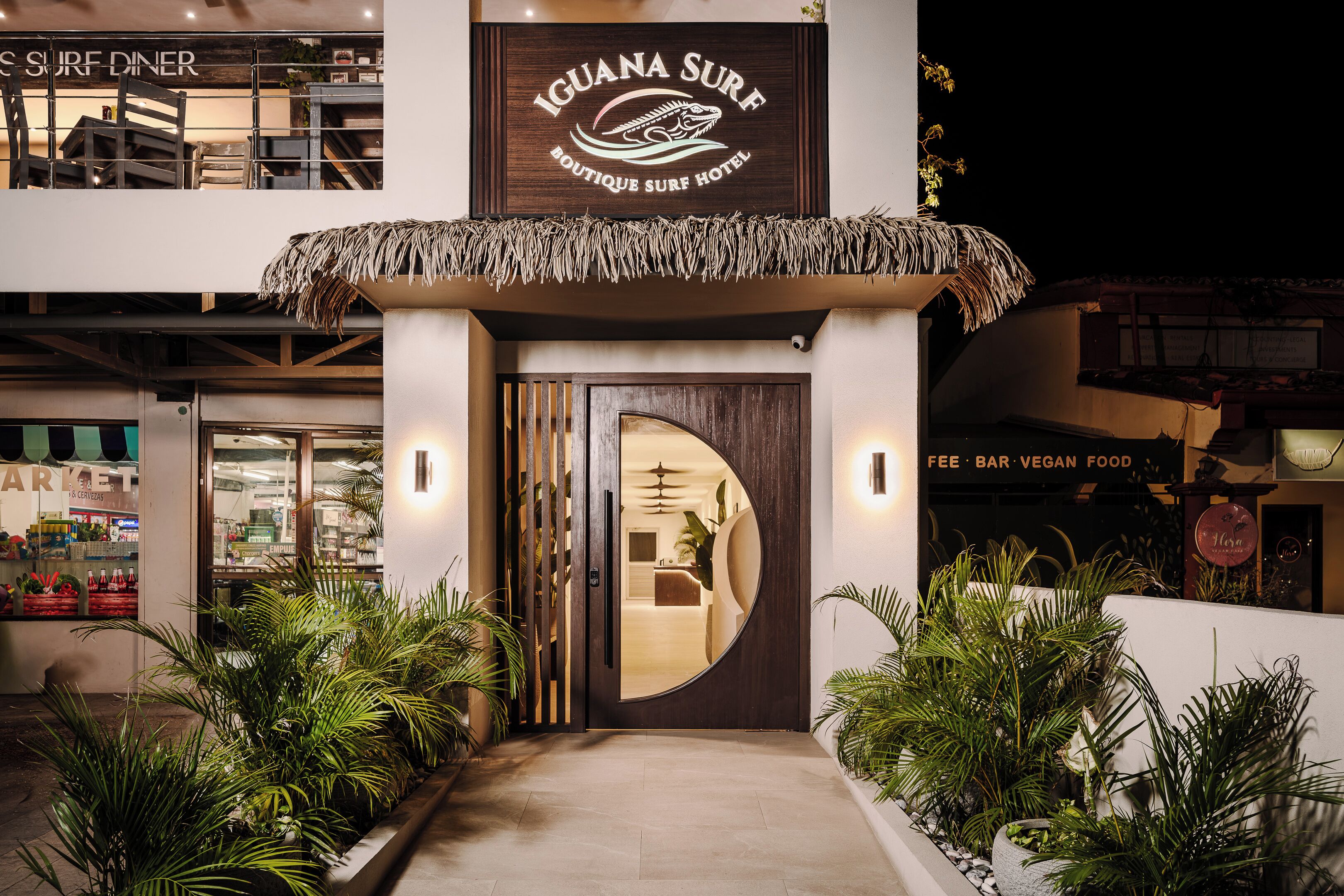The exterior of Iguana Surf Boutique Surf Hotel with a thatched roof entrance, a visible security camera, and a sign indicating a fee bar and vegan food options.