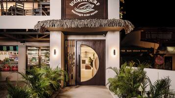 Exterior de l'Hotel Iguana Surf Boutique, amb entrada coberta de palla, càmera de seguretat visible i un cartell que indica bar amb cost i opcions de menjar vegan.