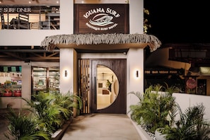 The exterior of Iguana Surf Boutique Surf Hotel with a thatched roof entrance, a visible security camera, and a sign indicating a fee bar and vegan food options. - Iguana Surf Boutique Hotel Tamarindo (Tamarindo)