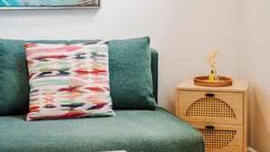 Une pièce avec un canapé vert, un coussin coloré, une table d’appoint en bois avec un panier, et un tableau accroché au mur.