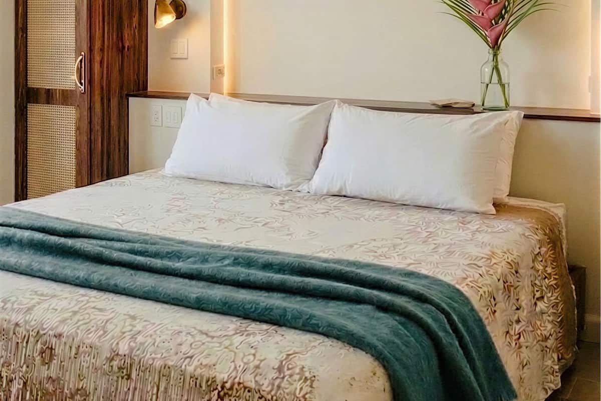 A hotel room with a bed, a wooden wardrobe, a wall-mounted light, and a vase with flowers.
