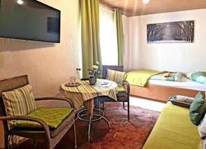 Ein Zimmer mit Bett, Tisch mit Stühlen, einem an der Wand montierten Fernseher und einem Wandbild.